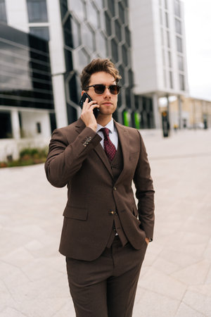 Vertical portrait of successful businessman in suit and sunglasses making call using mobile phone standing in city street on cloudy summer day, exuding style. Concept of business communicationの写真素材