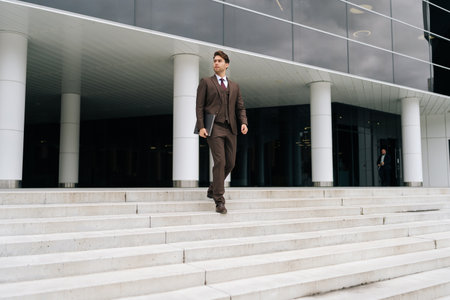 Stylish confident businessman in suit descending office building stairs with laptop in hand, showcasing professional demeanor. Successful business man walking down stairs of modern office building.の写真素材