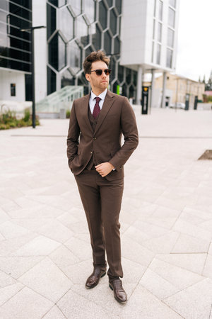 Full length vertical portrait of businessman in stylish brown suit standing in front of modern office building. Handsome serious business man in sunglasses posing outside holding hand in pocket.の写真素材