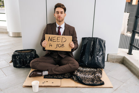 Distressed businessman in business attire looking at camera sitting on sidewalk with sign NEED JOB, illustrating difficulties of unemployment in modern society. Concept of young beggar in city.の写真素材