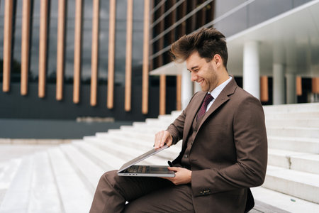Positive successful business man in fashion suit working outdoors on laptop and laughing at something sitting on steps by modern office building. Concept of business communication and technology.の写真素材