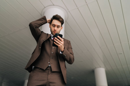 Low-angle view of businessman reacting to news looking on smartphone screen in standing by office building, showcasing pressures of business world. Concept of business communication.の写真素材