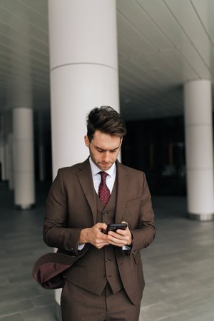 Vertical portrait of serious businessman in stylish suit standing in cityscape background, browsing smartphone app, immersed in digital world, looking to screen.の写真素材