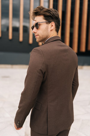 Vertical shot of confident businessman in fashion brown suit and sunglasses standing in city amidst modern architecture. Photogenic young business man posing outdoors alone enjoying summer day.の写真素材