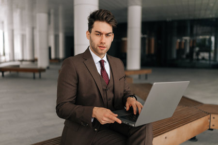 Portrait of bearded businessman in suit sitting on urban bench by modern office building, working on laptop, looking at camera with serious expression. Concept of business communication.の写真素材