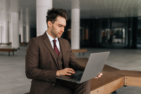 Serious focused businessman in stylish sharp brown suit working typing on laptop computer sitting outside by modern office building, showing determination and ambition in professional urban setting.の写真素材