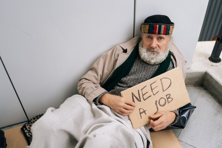 Portrait of sad elderly homeless man holding NEED JOB sign in city sidewalk, showcasing economic hardship and social inequality. Concept of poverty and vulnerability, evoking empathy and compassionの写真素材