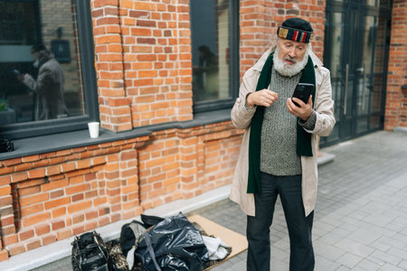Elderly bearded beggar man standing on urban street, using smartphone while holding conversation, with belongings lying on piece of cardboard nearby. Concept of poor unhappy retirement.の写真素材