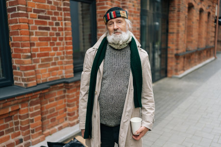 Portrait of tired older retired man wearing worn-out coat, holding paper cup while standing on city street in cold day, looking at camera with sadness facial expression.の写真素材
