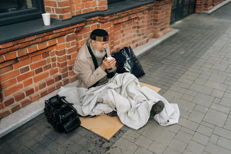 High-angle view of freezing aged homeless man sitting on city street, holding hot drink to warm hands, reflecting sadness and hope. Portrait of sad older tramp male with emotion of poverty and despairの写真素材