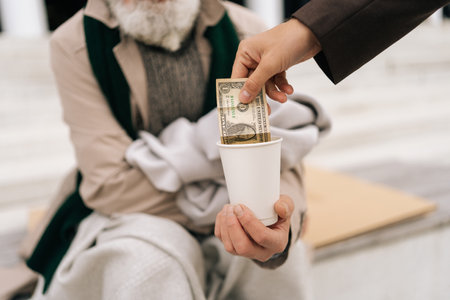 Closeup cropped shot of unrecognizable kind man giving paper money to senior homeless beggar male sitting on urban street, symbolizing hope and generosity in face of poverty and unemployment.の写真素材