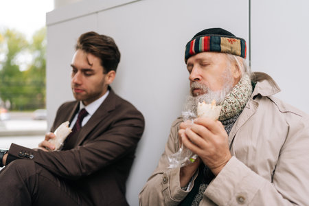 Kind young businessman in suit and elderly homeless man eating together sandwich on city street, bridging social divides with empathy and generosity, symbolizing charity and social inequality.の写真素材