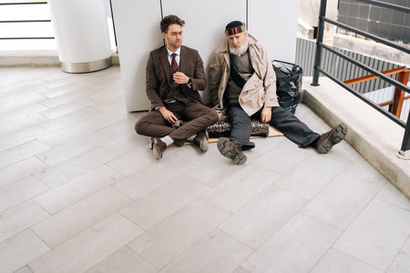 Contrasting image of chatting young businessman in suit with elderly homeless man sitting on cardboard, representing social and economic disparity. Concept of social issues and poverty.の写真素材
