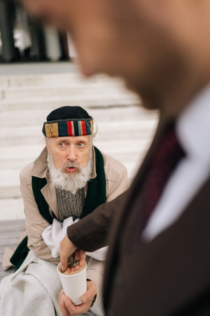 Vertical cropped shot of contrast between wealth and poverty as businessman donating coins to homeless beggar cup. Closeup hands of unrecognizable business man dropping coins into homeless beggar cup.の写真素材