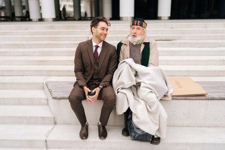 Portrait of cheerful business man listening and smiling as talking with elderly homeless male, demonstrating empathy and kindness in heartwarming moment of human interaction, sitting on urban steps.の写真素材