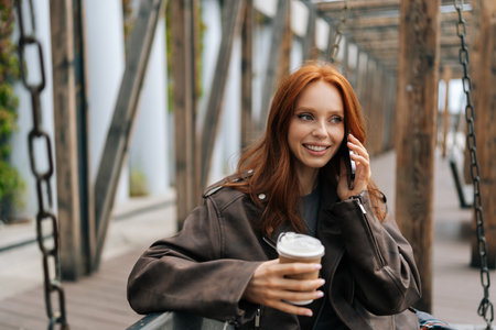 Waist up portrait of cheerful young woman with long red hair enjoying coffee on city swing, happily chatting on phone outdoors in city park on cloudy day. Happy red-haired female enjoy coffee in park.の写真素材