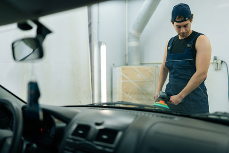 View through windshield of mechanic male in uniform polishing surface of hood of black car using orbital polisher in repair shop. Professional repairman in uniform polishing paintwork hood of car.の写真素材