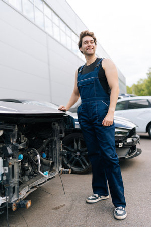 Full length vertical portrait of smiling mechanic in overalls standing by damaged car in repair shop, exuding positive attitude and showcasing expertise in fixing vehicles. Concept of car repairの写真素材