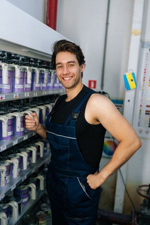 Vertical portrait of friendly auto painter male in uniform choosing appropriate color for car dyeing, smiling looking at camera, make choice from cans. Mechanic selection colors for car painting.の写真素材