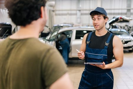 Male garage worker in uniform and unrecognizable man customer talking to discuss details of car repairs. Auto mechanic holding tablet and writes repair list for client in auto repair workshop.の写真素材