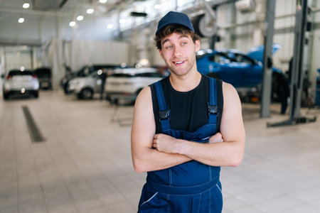Portrait of cheerful male car repairman in overalls standing in workshop or garage, confidently looking away background of cars under repair. Concept of car service, repair and maintenance.の写真素材
