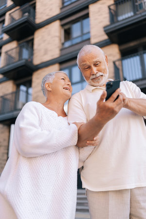Low-angle view vertical portrait of cheerful beautiful senior couple with gray-haired in white shirts standing on city street, happily browsing using smartphone, radiating love and joy.の写真素材
