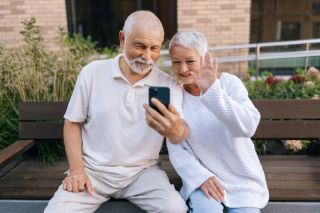Portrait of happy retired family couple using mobile phone for video call together, talking to relations, getting good news, having fun, laughing, smiling looking to screen, sitting on urban benchの写真素材