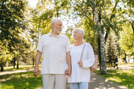 Portrait of beautiful senior couple loving looking at each other, holding hands, enjoying leisurely walk in summer park with lush green on background. Concept of happy retirement life.の写真素材