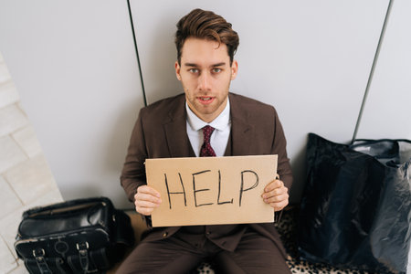 High-angle view of homeless man wearing suit and tie sitting on street with cardboard sign asking for help. Unhappy young man seeking assistance after losing everything.の写真素材