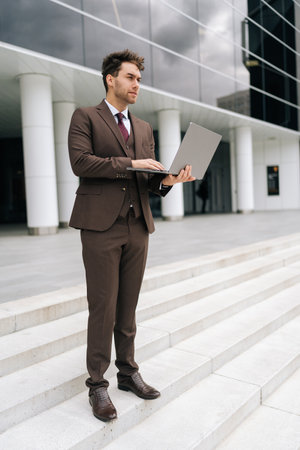 Vertical portrait of businessman in suit standing outside modern office building working typing on laptop with pensive expression. Handsome business man working remote online standing urban setting.の写真素材