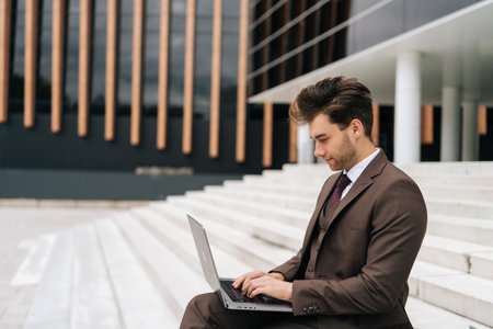 Side view of ambitious young business man in fashion suit focused working on laptop computer sitting on steps by modern city office building, showcasing determination in modern business world.の写真素材
