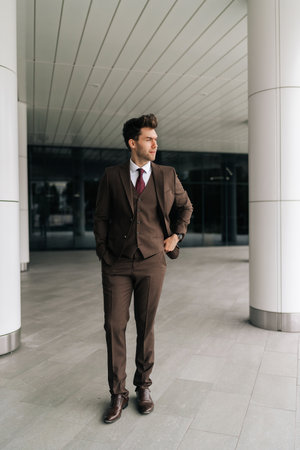 Full length vertical portrait of confident successful businessman in suit walking around by modern office building, conveying sense of purpose and determination, looking away with serious expression,の写真素材