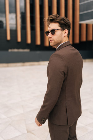 Vertical shot of serious confident businessman wearing brown suit and sunglasses standing outside of modern office building, looking over shoulder. Photogenic business man posing outdoors alone.の写真素材