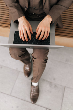 Top view of business man in brown suit is sitting on bench, typing on laptop, working remotely, taking advantage of opportunity to work outside on fresh air. Concept of business communication.の写真素材