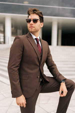 Vertical portrait of serious confident businessman in stylish suit and sunglasses posing outdoors, exuding success and professionalism, city architecture complementing modern urban look.の写真素材