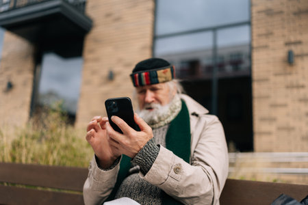 Senior man sitting on urban bench, using smartphone, checking messages or browsing internet, highlighting intersection of technology and modern society.の写真素材