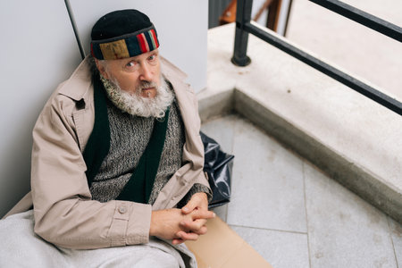 Portrait of unhappy elderly homeless man with beard sitting lying on sidewalk with belongings, seeking assistance. looking at camera with sad expression. Concept of poverty and vulnerability.の写真素材