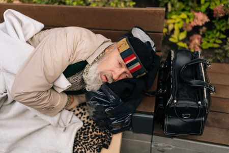 High-angle view of tired sad aged homeless man lying on bench outside, seeking refuge from elements. Sad beggar aged male bundled up in warm clothing lying with bag of belongings.の写真素材
