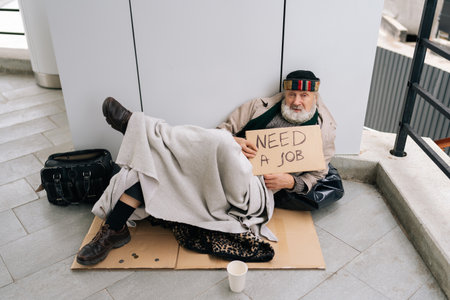 Full length portrait of alone despair elderly homeless man with sign seeking employment on urban street, appearing distressed and hopeless amid economic downturn. Concept of poor unhappy retirement.の写真素材