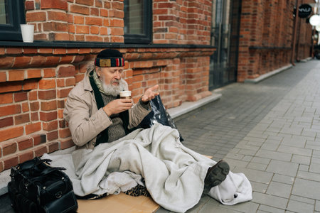 Portrait of frozen senior man with grey beard, dressed in beige coat, enjoying hot tea from paper cup while warming up sitting on urban street during cold autumn. Concept of poor unhappy retirement.の写真素材