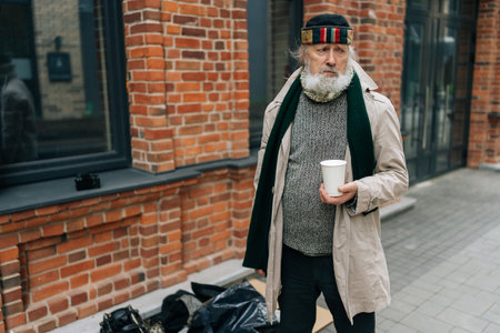 Portrait of senior adult homeless man with lengthy white beard and poor attire, clutching white paper cup while standing by his possessions on city sidewalk. Concept of poverty and despair.の写真素材