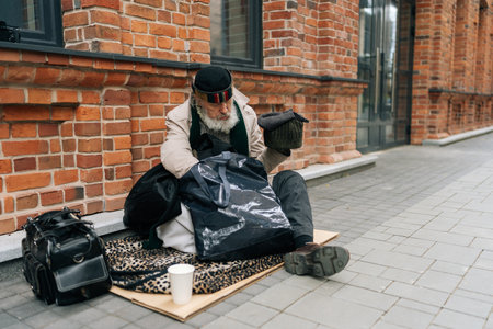 Elderly homeless man in hat sitting outside in cold, holding out cup for help in urban street, reflecting issue of homelessness. Homeless retired male huddled on city street holding legs for warmth.の写真素材