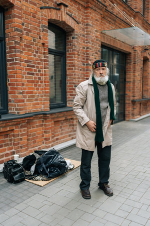 Full length vertical portrait of elderly tramp man in coat and scarf standing on street beside meager possessions, confronting hardship and destitution, looking at camera. Concept of poor retirement.の写真素材