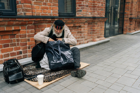 Wide shot of homeless aged man with long white beard searching through belongings on cold day in city, reflecting social issue of poverty and homelessness. Concept of a poor unhappy elderly.の写真素材