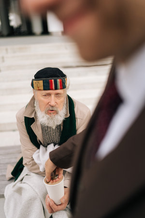Vertical shot of contrast between wealth and poverty as businessman donating coins to homeless beggar cup. Closeup hands of unrecognizable business man dropping coins into homeless beggar cup.の写真素材