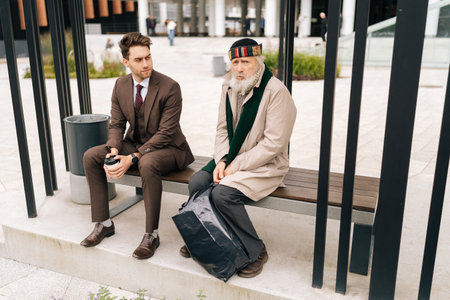 Handsome businessman in suit holding coffee cup, talking and listening to sad senior adult homeless man while sitting on urban bench at bus stop. Concept of helping older tramp people.の写真素材