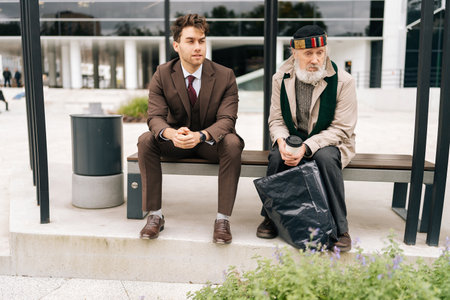 Wide shot of chatting successful businessman and old homeless man sitting on bench, representing growing gap between rich and poor in modern society. Concept of social issues and economic disparity.の写真素材