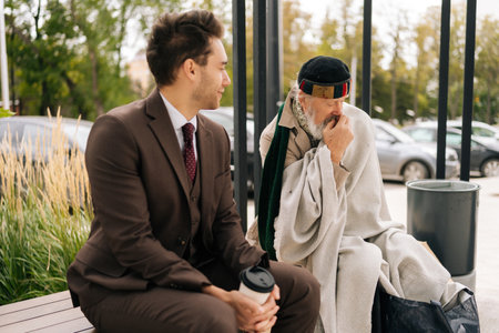 Compassionate entrepreneur with coffee engaging in conversation with homeless senior citizen. Kind businessman in suit listening with empathy to homeless elderly man talking sitting on urban bench.の写真素材