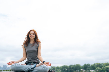 Portrait of smiling young woman in casual clothes sitting in lotus position on wooden platform by river, practicing yoga and meditating. Serene female enjoying peace and tranquility of nature aloneの写真素材