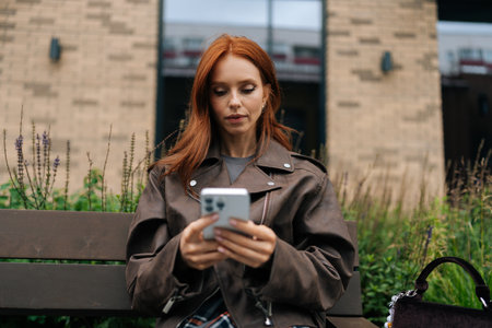 Low-angle portrait of focused young woman with red hair concentrating on smartphone sitting on bench outside. Redhead female in brown leather jacket using smartphone outdoors, enjoying free time.の写真素材